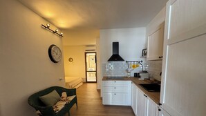 A compact kitchen with white cabinets, a wooden countertop, and a black range hood. A green chair is placed in the living area, and a clock is visible on the wall.