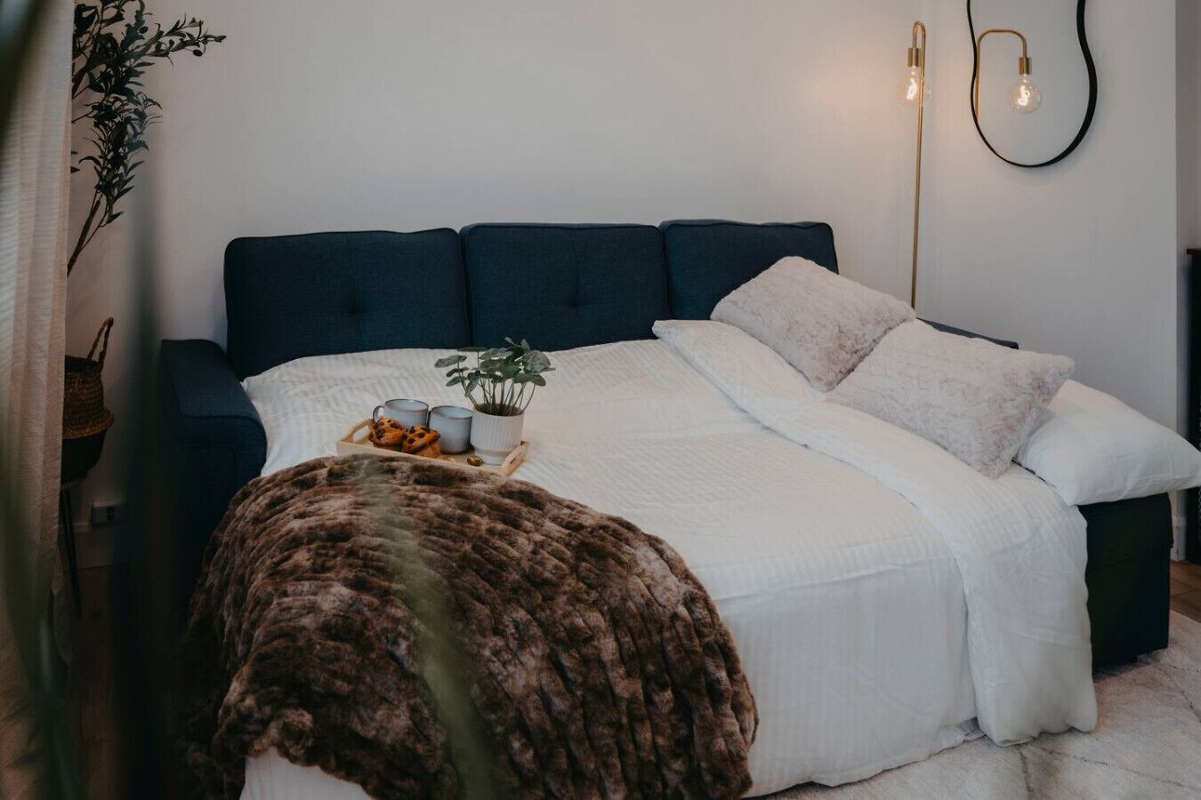 A bedroom with a dark blue headboard, white bedding, a fluffy brown throw, a tray with cookies, and a potted plant.