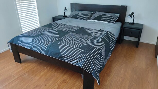 Een bed met een sprei met geometrisch patroon, twee nachtkastjes met lampen, en een raam met jaloezieën.