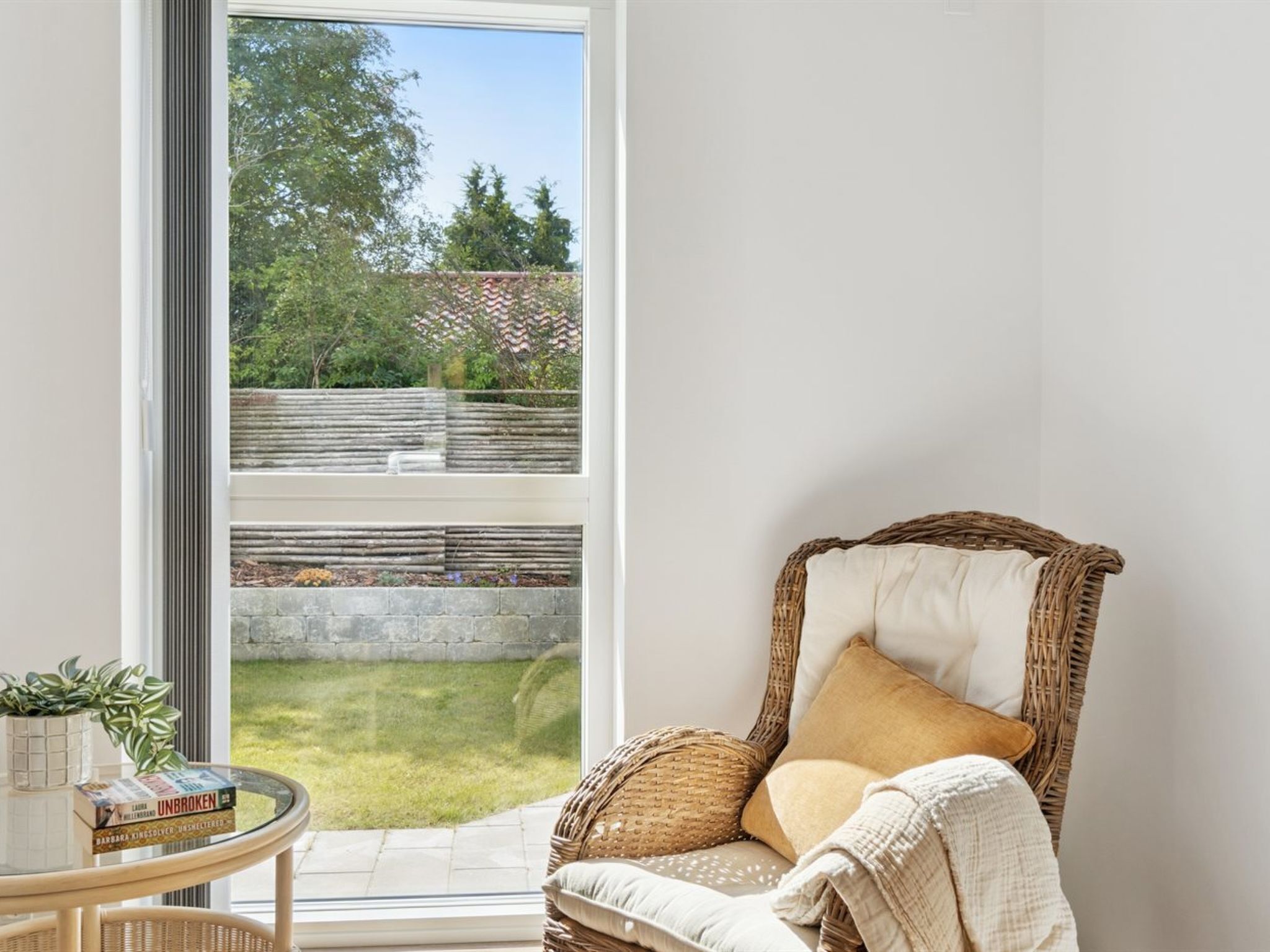 A corner room with a wicker chair, a small table with books and a plant, and a large window overlooking a garden.