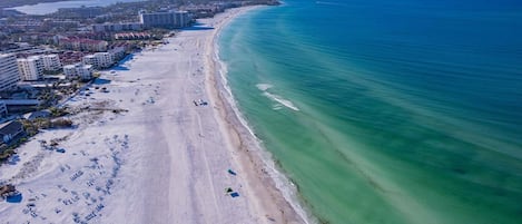 An aerial view of a beach with buildings along the shore, clear blue water, and a sandy beach.