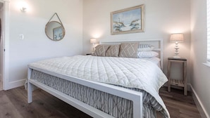 Un dormitorio con cama, dos mesitas de noche, un espejo montado en la pared, un cuadro enmarco y una ventana con persianas.