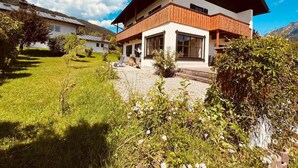 A modern two-story house with a wooden exterior and large windows, surrounded by a well-maintained garden and a mountainous backdrop.