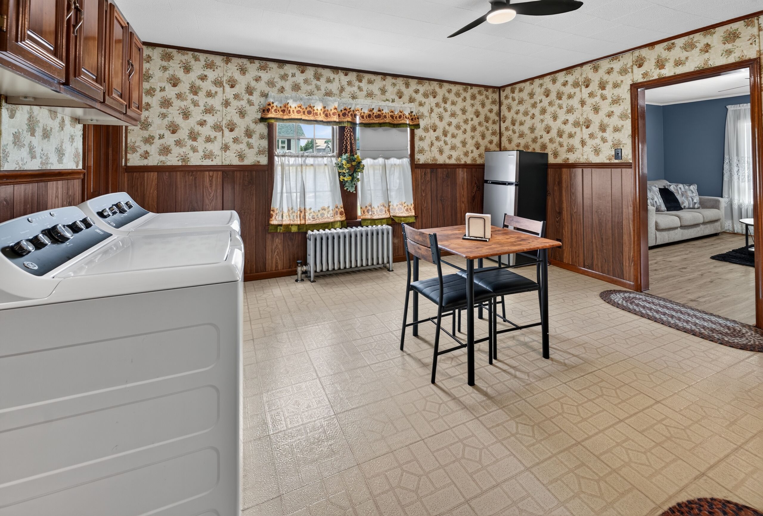Een compacte kamer met een kitchenette, een eettafel met stoelen, een radiator en een raam met gordijnen.