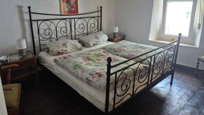 Un dormitorio con estructura de cama metálica, ropa de cama floral, una mesita de noche con lámpara y una ventana con cortinas.