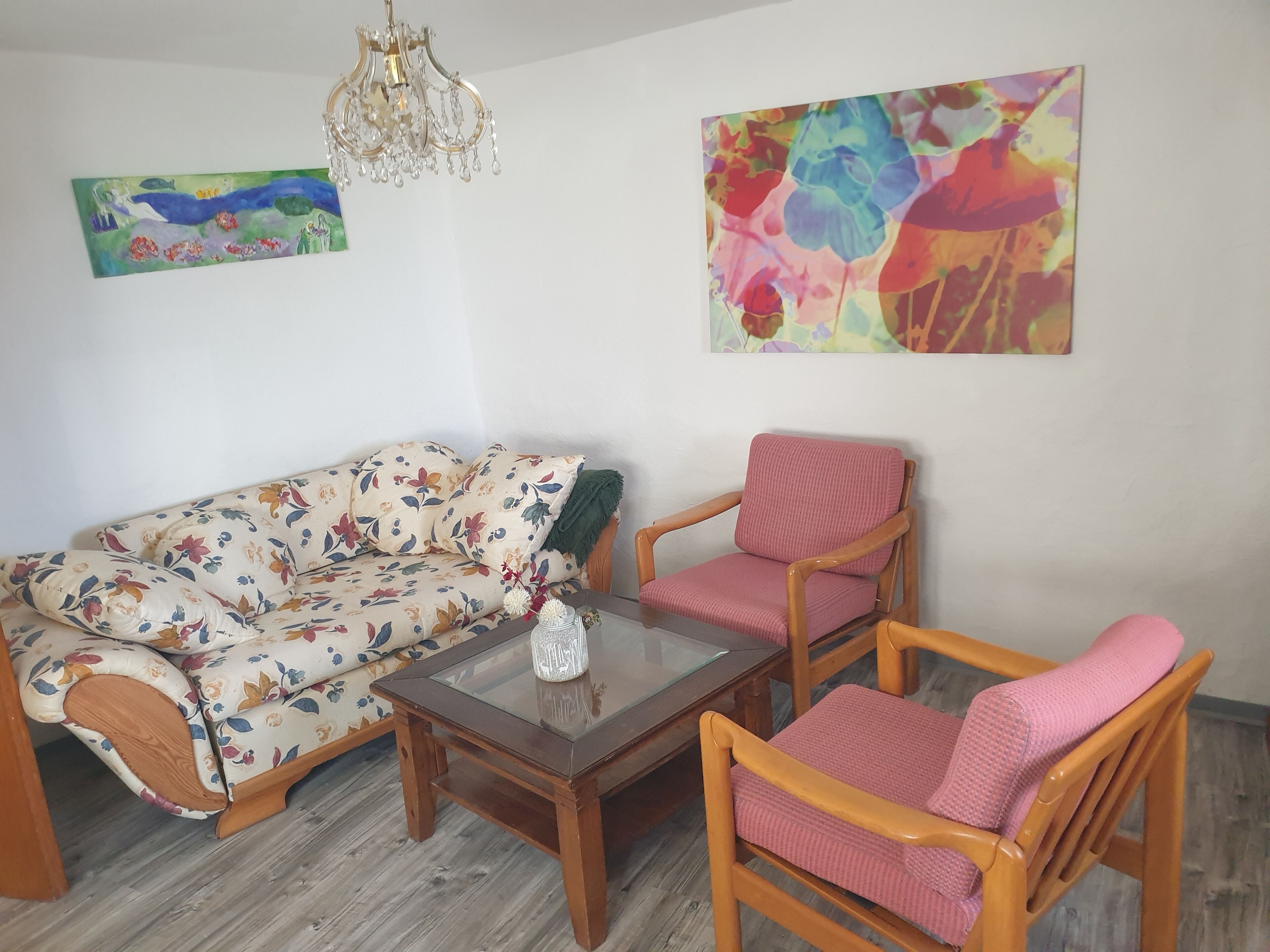 Sala de estar com sofá floral, poltronas de madeira, mesa de centro de vidro e um candelabro.