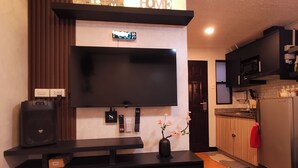 A modern living room with a flat-screen TV, a black entertainment unit, a wooden panel wall, a welcome sign, a digital clock, and a kitchen area with cabinets and appliances. - Stay Near Philippine Arena Chiara’s Stay Inn—Clean, Calm, and Close to It All (Marilao)