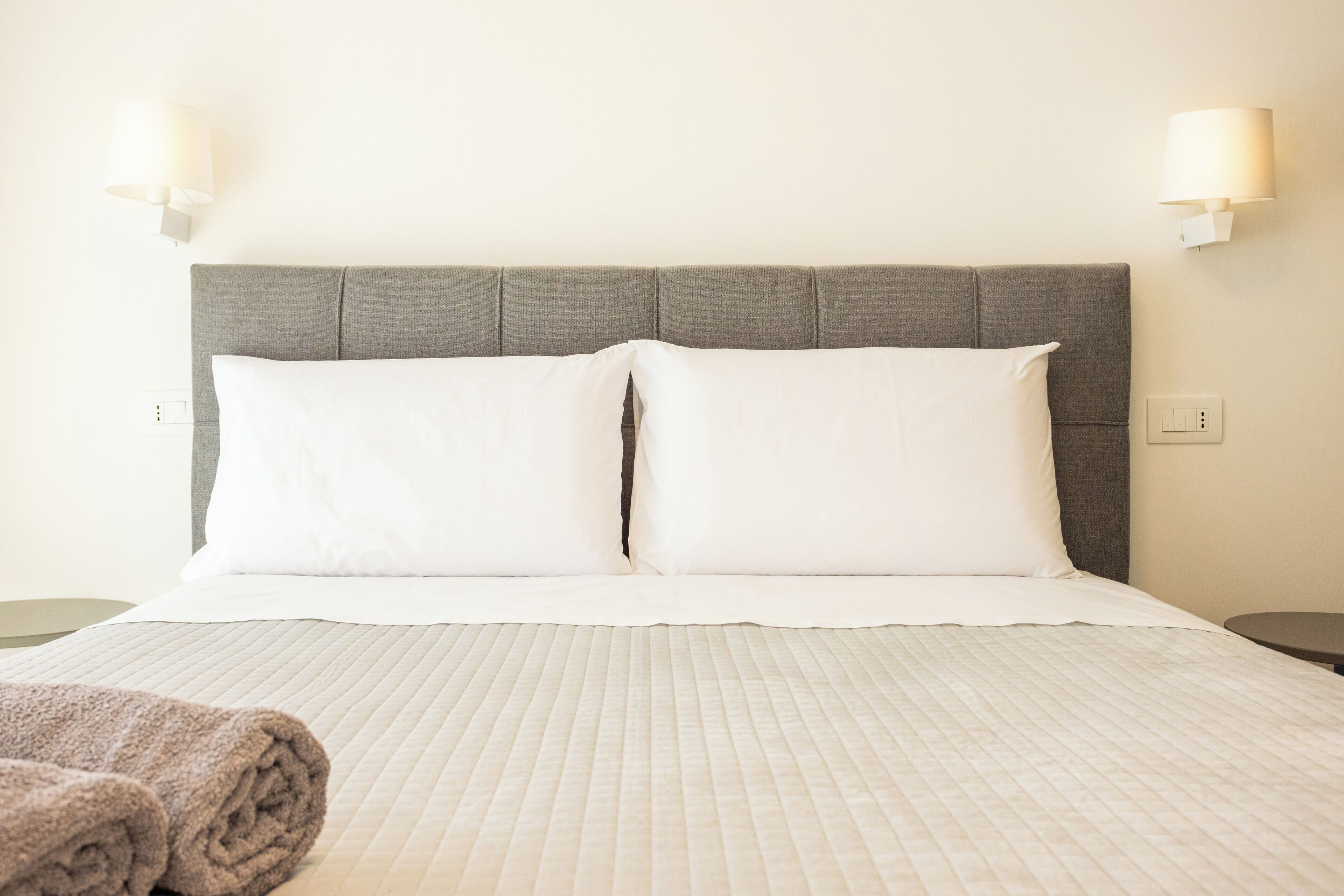 A neatly made bed with a grey headboard, white pillows, and a folded towel on the side.