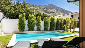 Une piscine entourée de chaises longues, bordée d’un mur blanc et de verdure, avec une chaîne de montagnes en toile de fond.
