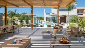 A covered outdoor area with lounge chairs, a pool, and a modern building in the background. - Lanik Luxury Pool Villa (Αφάντου)