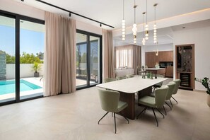 A modern dining area with a large table and chairs, a kitchen in the background, and a pool outside.