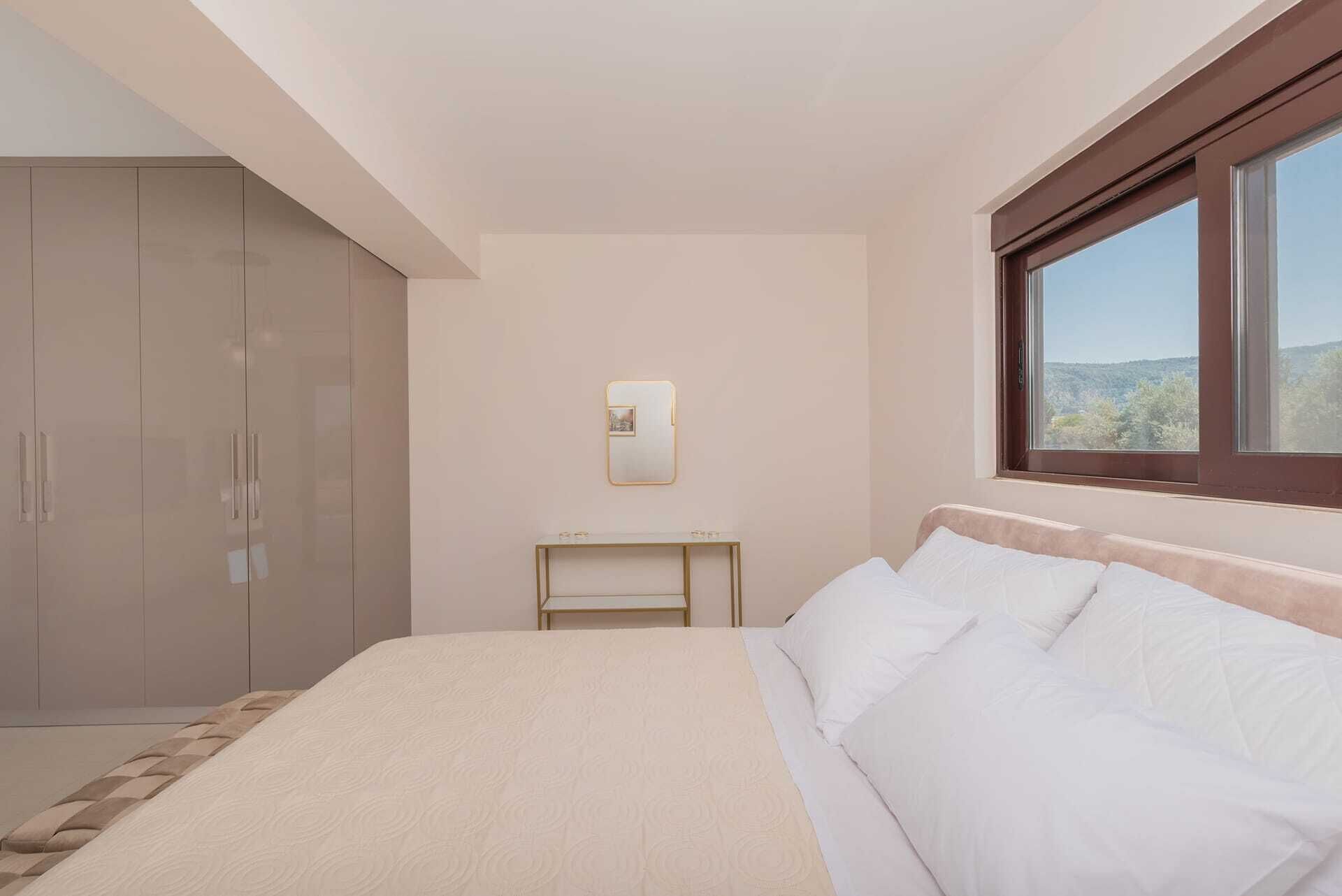 A bedroom with a bed, a wardrobe, a bedside table, a mirror, and a window with a view.