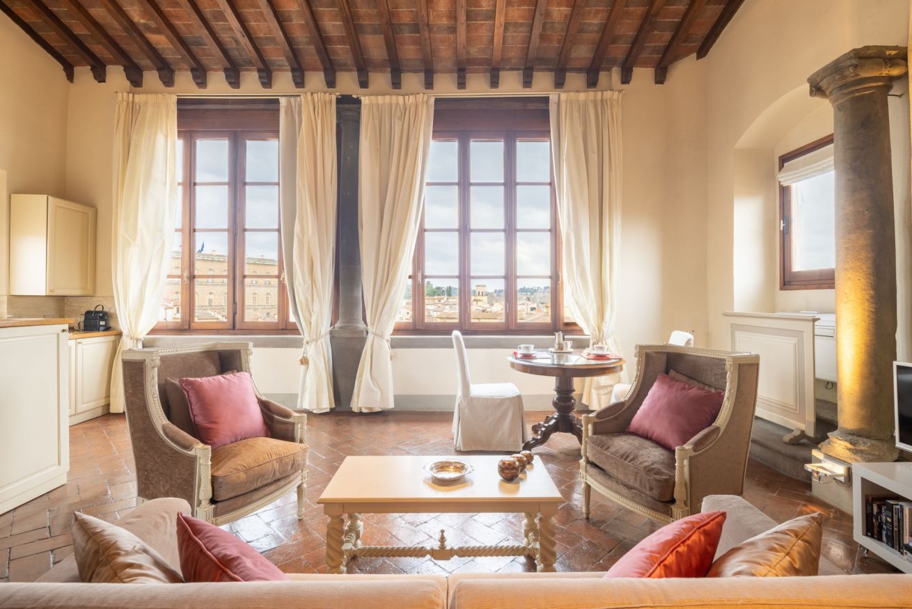A spacious living room with a high ceiling, two large windows with curtains, a round table, and two armchairs.