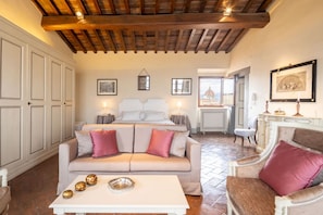 A cozy room with a bed, a sofa, a coffee table, and a chair. There are framed pictures on the walls and a window with a view.