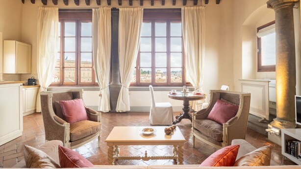 A spacious living room with a high ceiling, two large windows with curtains, a round table, and two armchairs. - Tower View Apartment (Firenze)