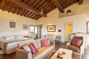 A spacious room with a large bed, a sofa, a chair, and a small table. There is a wooden ceiling and a door leading to another room.