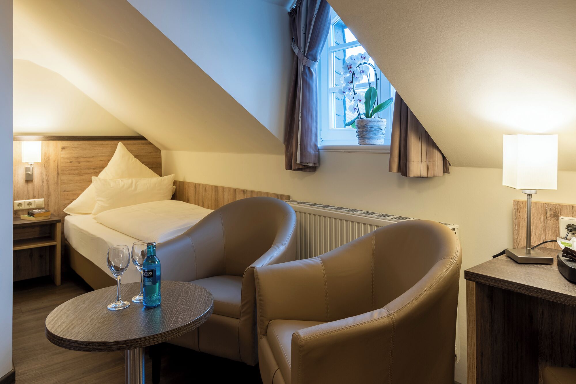 Habitació d'hotel acollidora amb un llit, dues butaques, una taula petita amb begudes, una finestra amb cortines i un radiador.