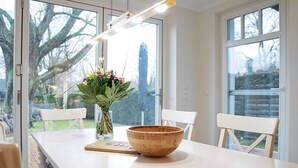 Un coin repas avec une table blanche, deux chaises blanches, un bol en bois, un vase avec des fleurs et une planche en bois.