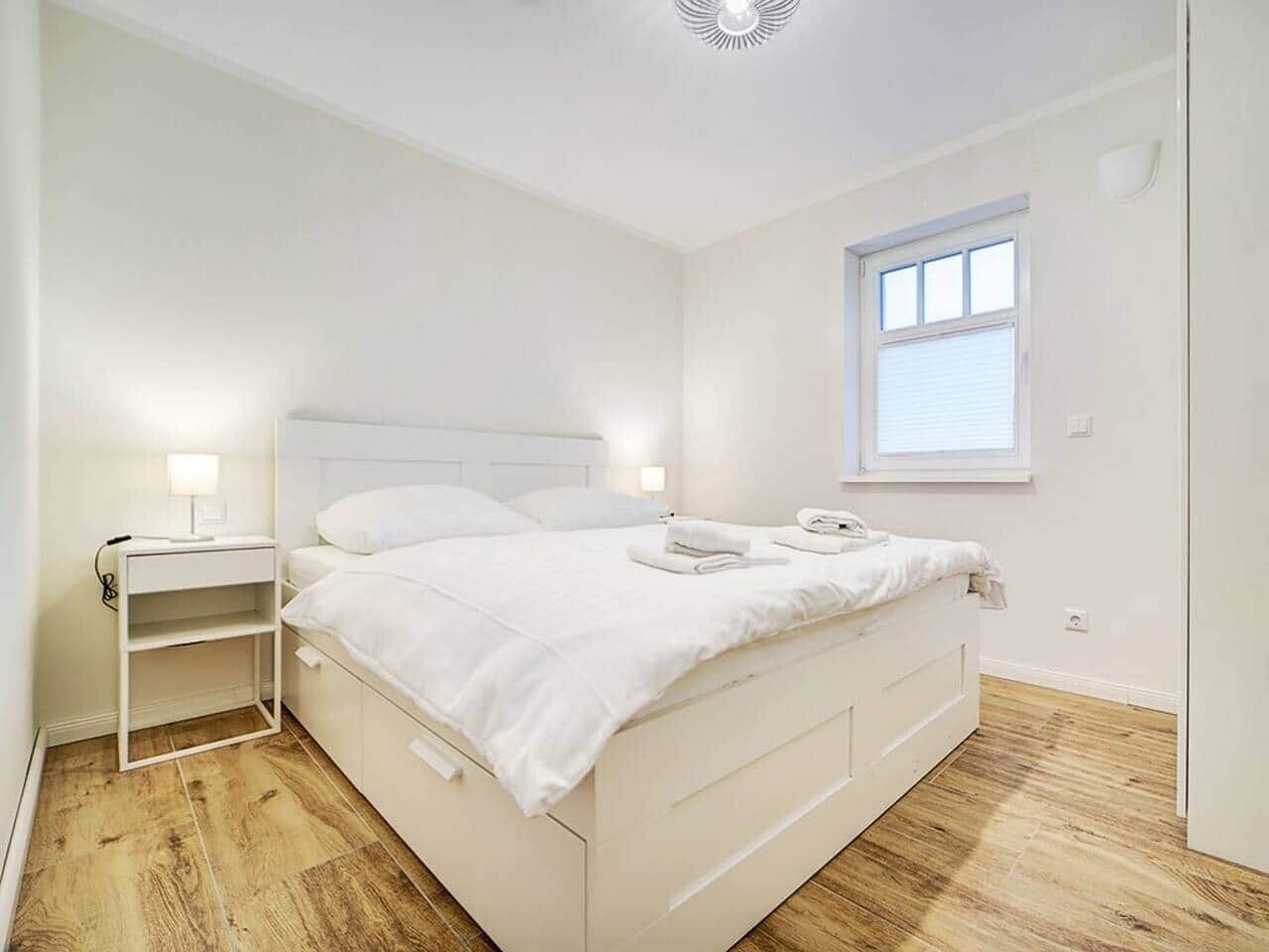 Un dormitorio blanco con cama, mesitas de noche y ventana.