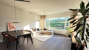 A modern living room with a dining area, large windows, and a view of a green landscape.