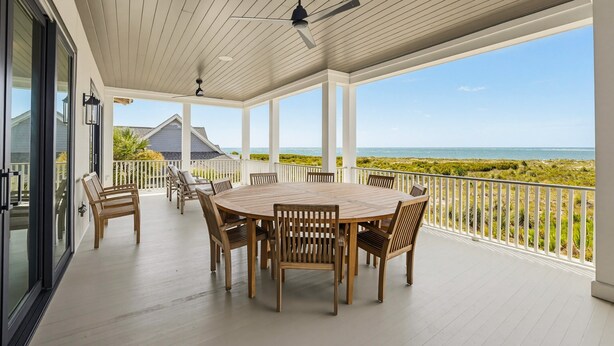 A covered outdoor patio with a round wooden table and chairs, a ceiling fan, and a view of the ocean.