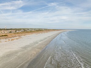 A coastal beach with a sandy shoreline, a clear sky, and a distant view of a town with houses and a water tower.