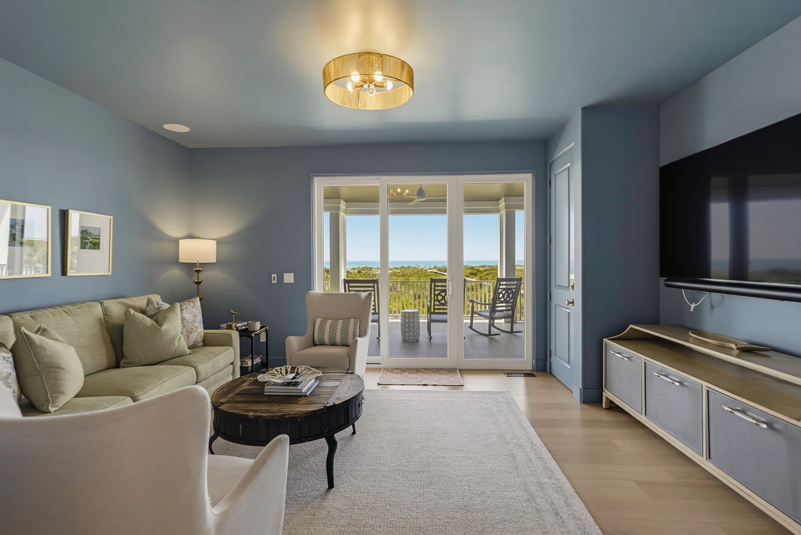 A living room with a sofa, coffee table, and a television. There is a sliding glass door leading to a balcony with outdoor furniture.