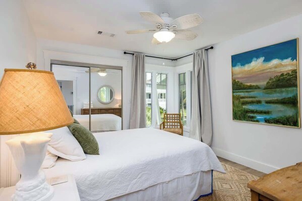 Un dormitorio con cama, lámpara, ventilador de techo, un cuadro y vistas al exterior.