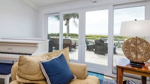 A room with a piano, a sofa with cushions, a wicker table, and a view of the beach through large glass doors. - 715 Winter Trout Road (Fripp Island)
