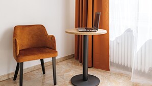 Quarto moderno com uma única cadeira marrom, uma pequena mesa redonda com um laptop e uma grande janela com cortinas leves.
