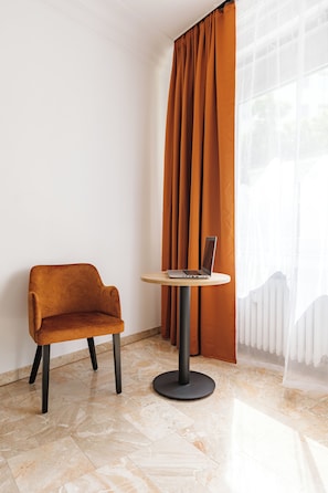 Ein modernes Zimmer mit einem einzelnen braunen Sessel, einem kleinen runden Tisch mit einem Laptop und einem großen Fenster mit transparenten Vorhängen.