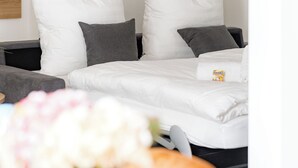 Quarto de hotel com cama, almofadas, mesa de cabeceira com um cesto de flores e uma pequena mesa com um prato e uma xícara.