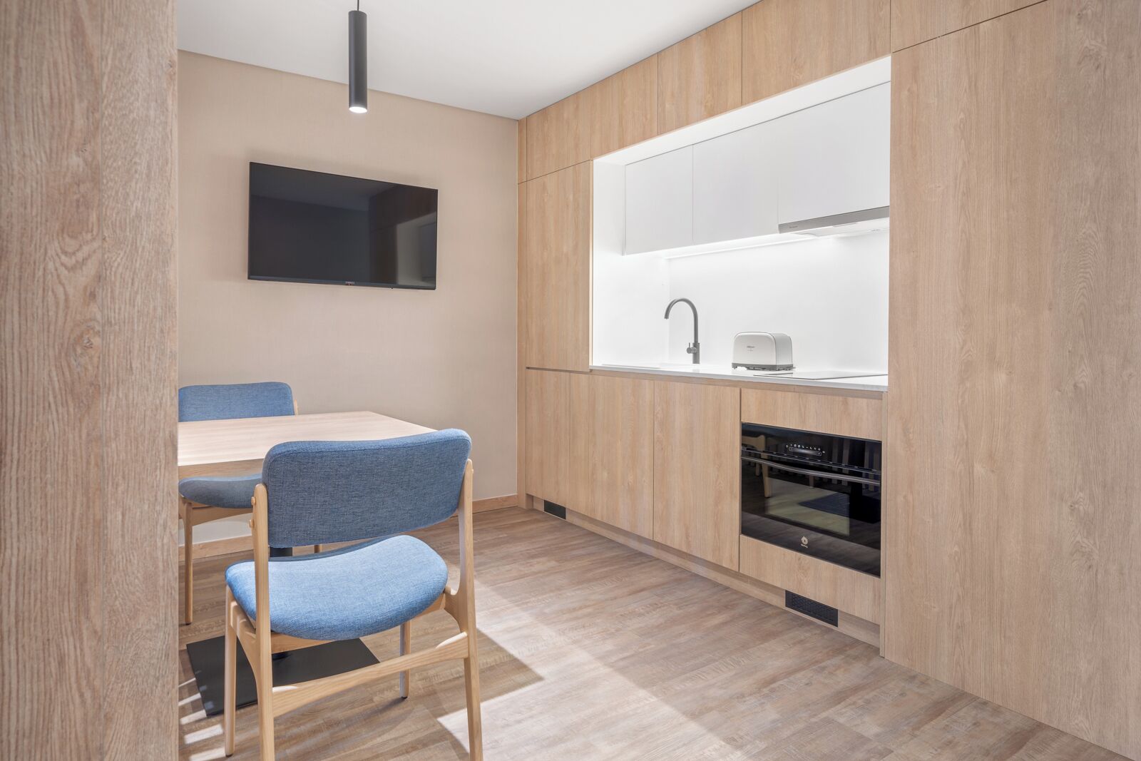 A modern kitchen with wooden cabinets, a built-in oven, a sink, and a dining area with a table and chairs.