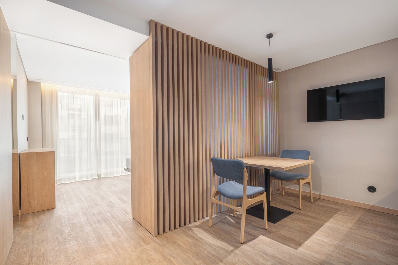 Un espace repas moderne avec un séparateur en bois, un téléviseur fixé au mur et une table en bois accompagnée de deux chaises.