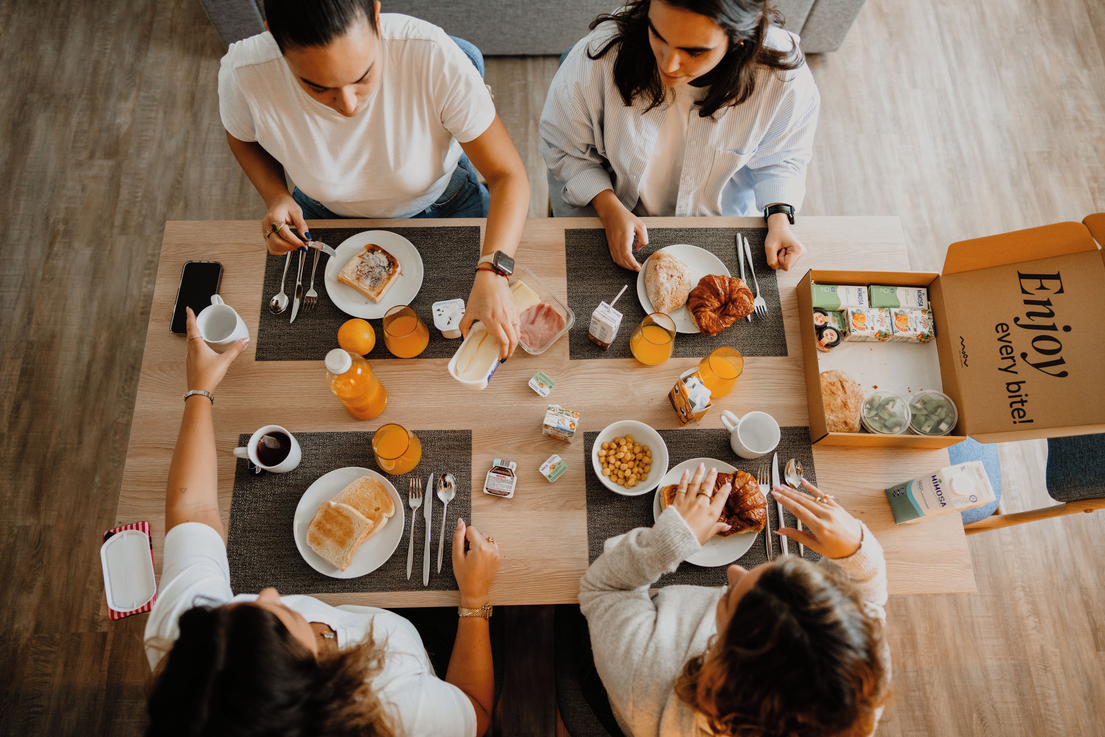 A group of people seated around a table with breakfast items, a box of food, and a smartphone.