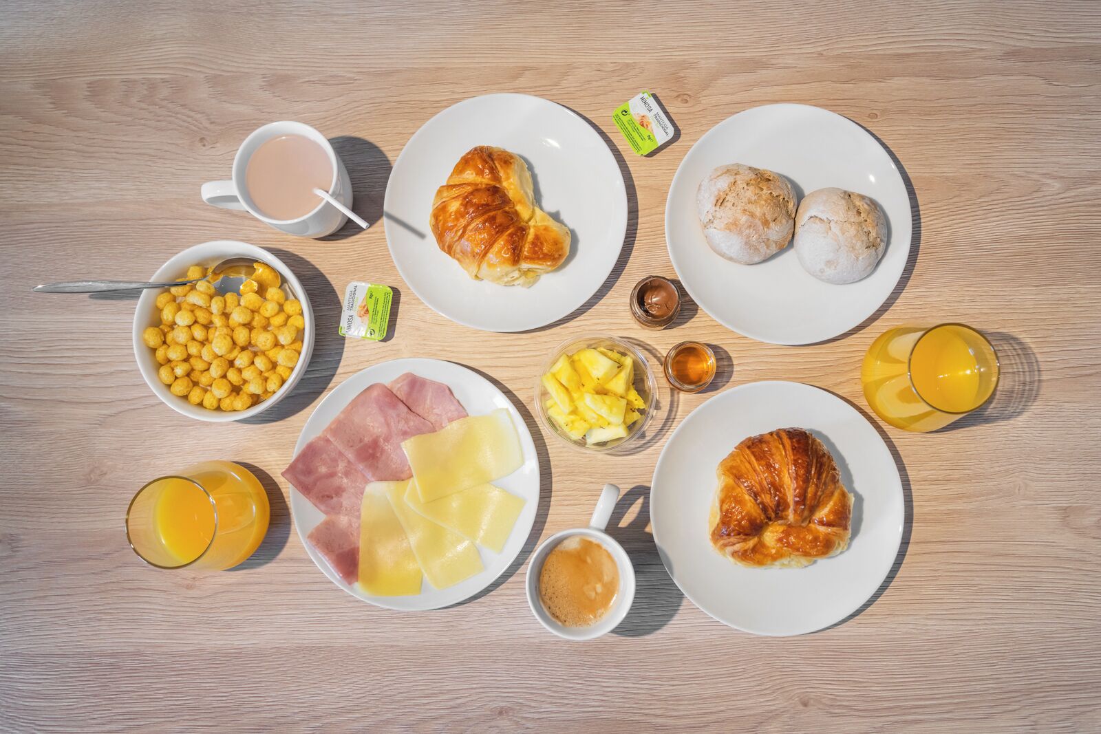A breakfast spread on a wooden table including croissants, ham, cheese, cereal, fruit, and beverages.