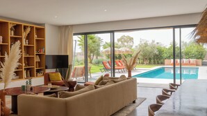 Un salon moderne avec un grand canapé, un téléviseur et une bibliothèque. On peut apercevoir une piscine à travers les grandes fenêtres.