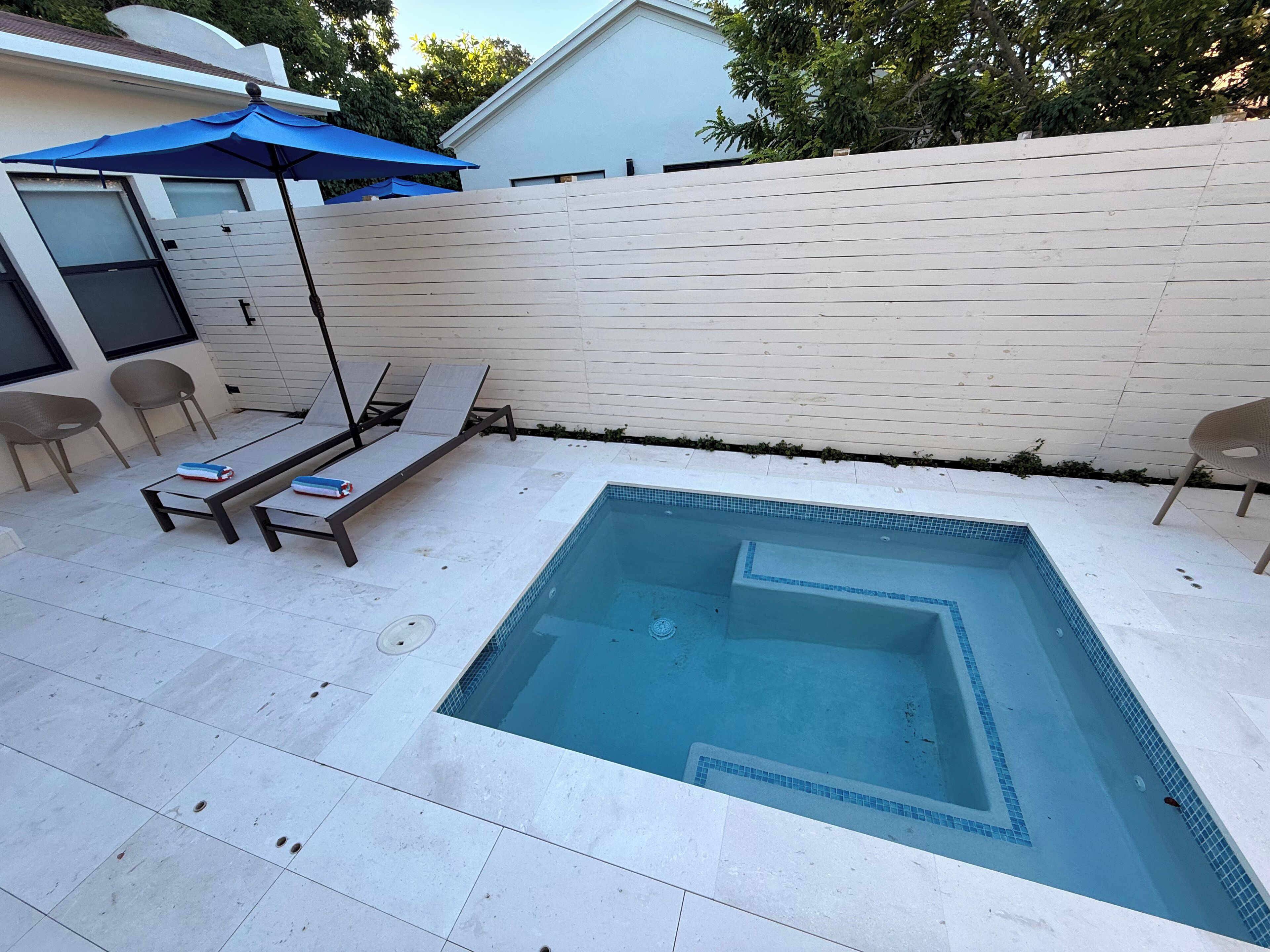 A rectangular swimming pool with a tiled border, surrounded by a white fence and a white wall. There are lounge chairs and a blue umbrella near the pool.