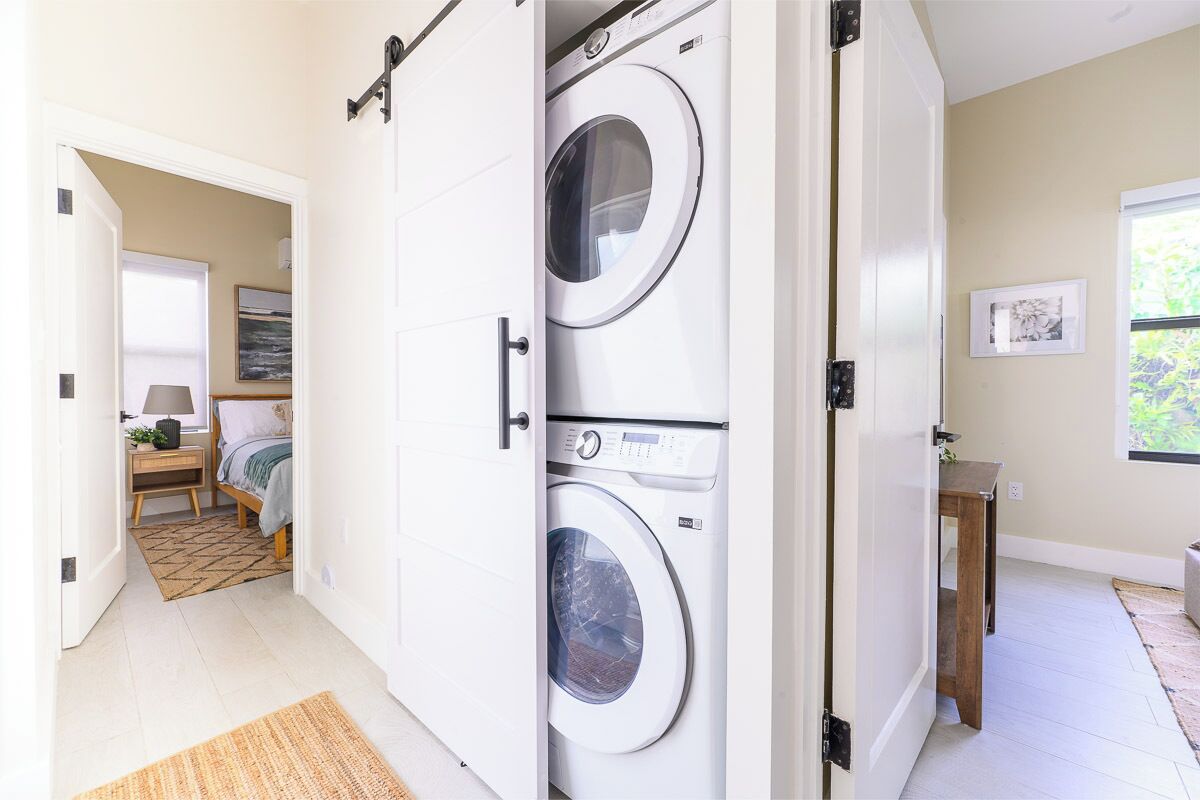 A laundry room with a front-loading washer and dryer, a wooden nightstand with a lamp, and a bed with a patterned rug.