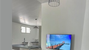 A modern living room with a glass-top coffee table, a flat-screen TV mounted on the wall, and a kitchen area with a bar counter and stools. - NEW: THE IVORY 3-BEDROOM VILLA (Gros Morne)