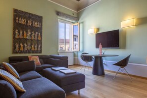 Un salon moderne avec un canapé, un pouf et une table à manger avec des chaises. Un grand tableau orne le mur et un téléviseur à écran plat est fixé au-dessus de la table.