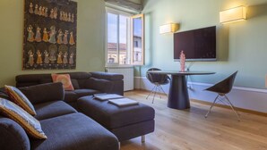 Ein modernes Wohnzimmer mit einer Couch, Hocker und einem Esstisch mit Stühlen. An der Wand hängt ein großes Gemälde und über dem Tisch ist ein Flachbildfernseher angebracht.