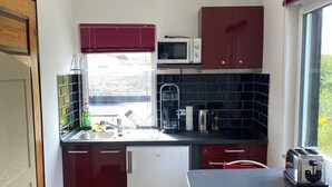 Cocina compacta con refrigerador, microondas y tostadora. Hay una ventana con persianas, un lavamanos y una pequeña zona de comedor con sillas.