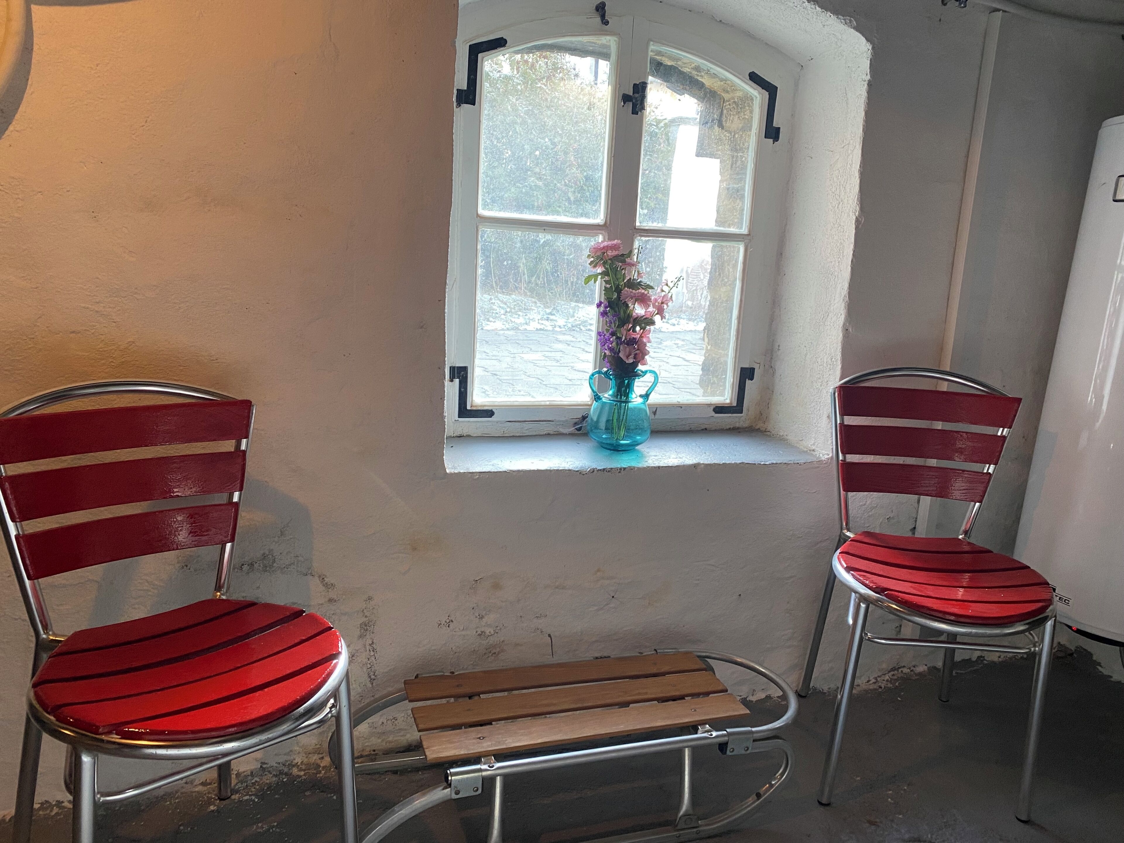 Zwei rote Stühle mit Metallgestell und eine Holzbank vor einem Fenster mit einem Blumenstrauß.