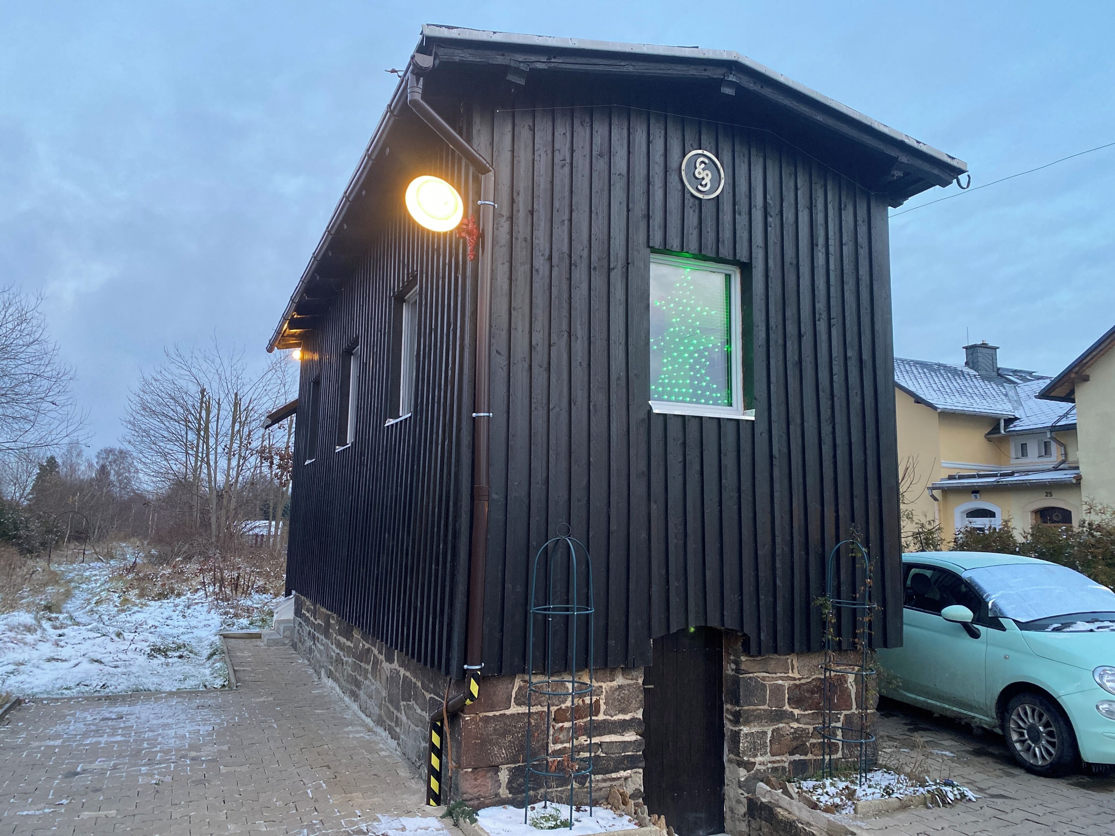 Une cabane en bois avec une fenêtre illuminée de lumières vertes, des fondations en briques et une voiture garée à proximité.
