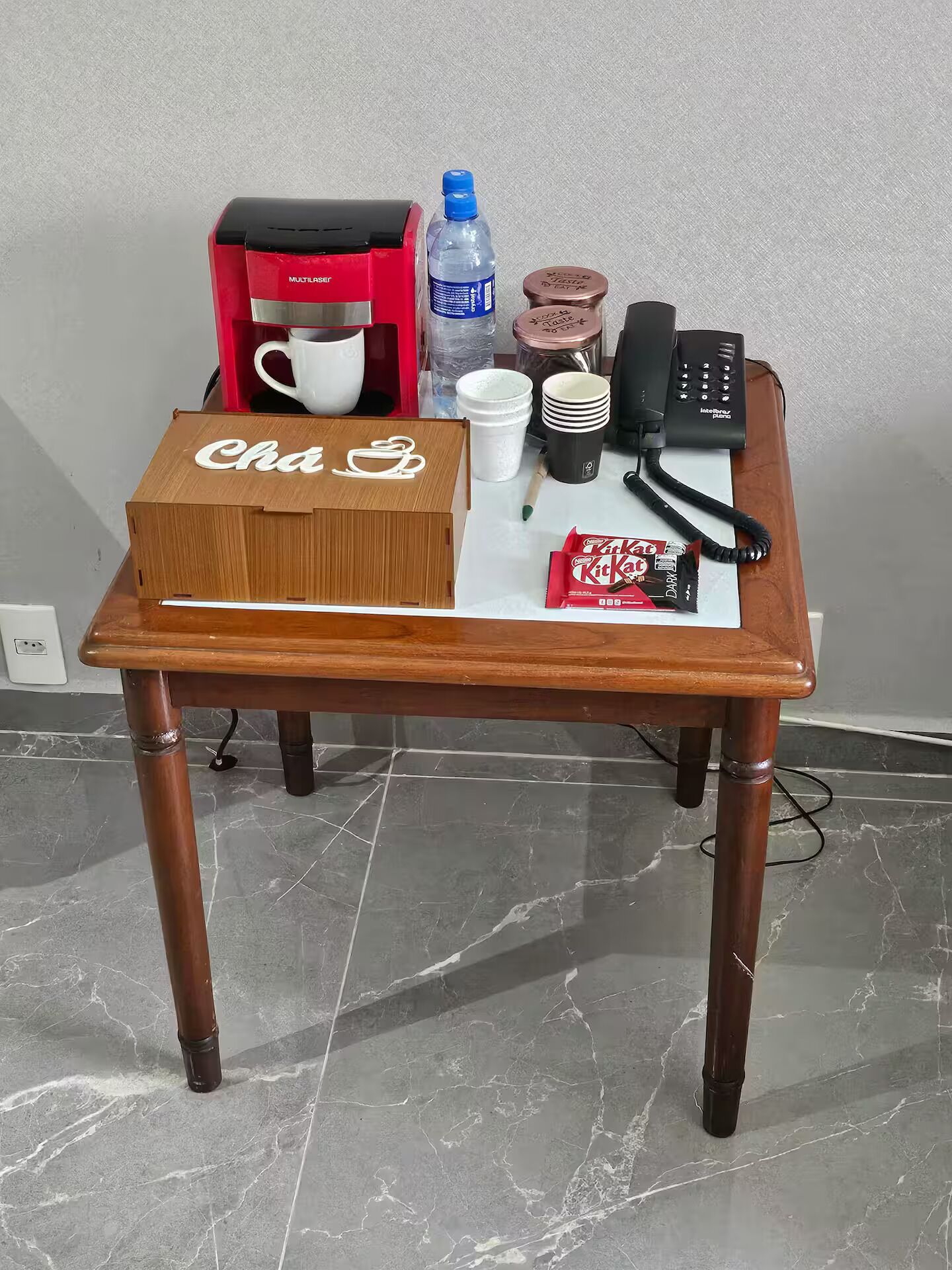 Ein Holztisch mit einer Kiste mit dem Aufdruck 'Cha & Go', einer Kaffeemaschine, Tassen, einer Wasserflasche, einem Einmachglas, einem Telefon und KitKat-Riegeln.