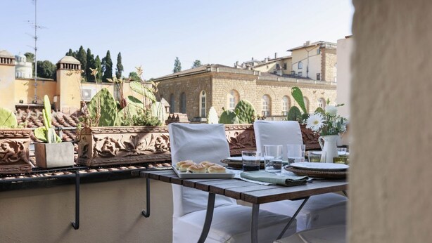 An outdoor dining area with a table set for two, surrounded by white chairs. There are potted plants and a view of historic buildings in the background. - Pitti Secret Rooftop (Firenze)