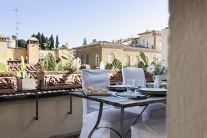 An outdoor dining area with a table set for two, surrounded by white chairs. There are potted plants and a view of historic buildings in the background.
