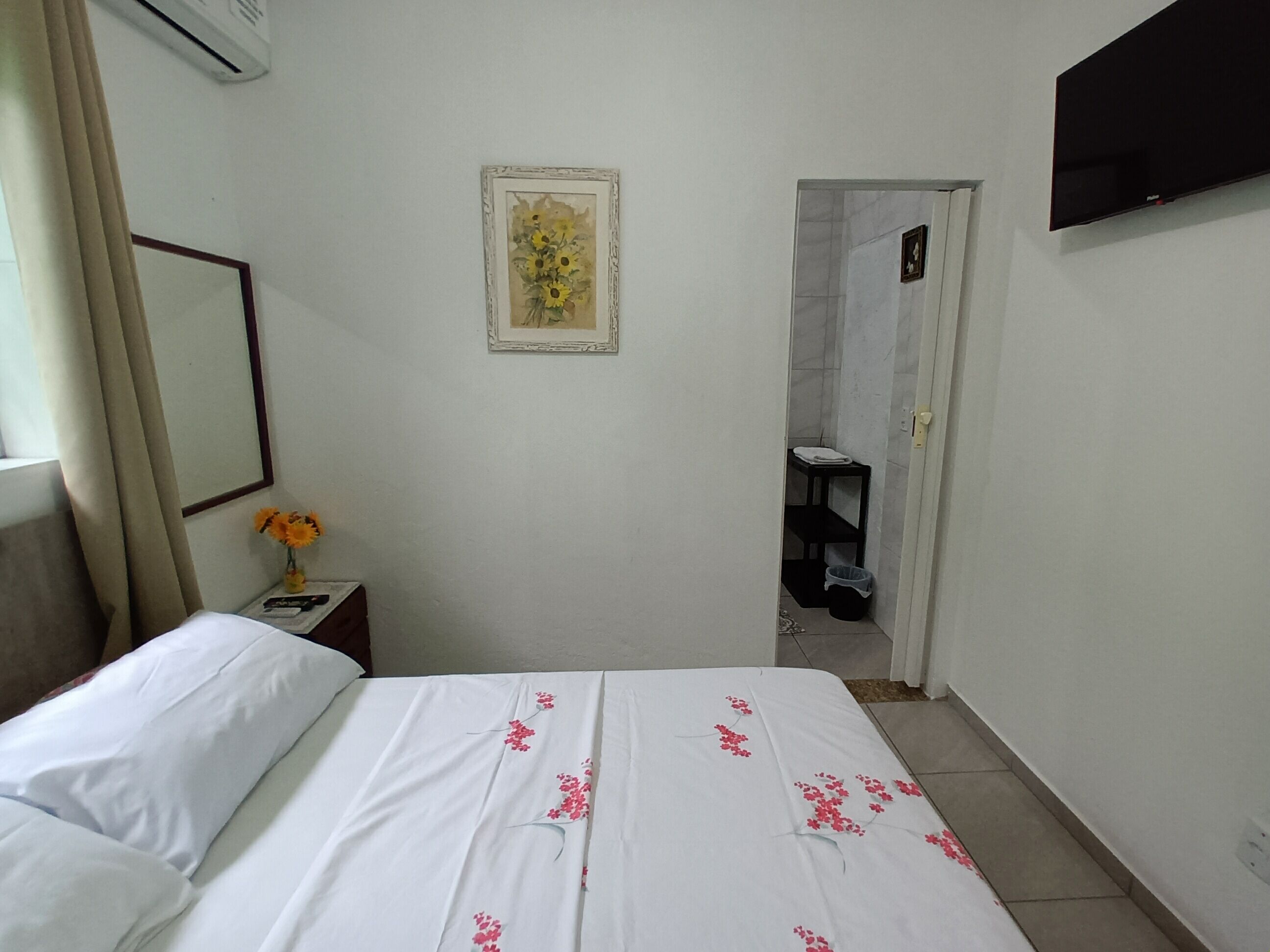 Un dormitorio con cama, una mesita con un jarrón de flores, un cuadro en la pared y un televisor montado en la pared.