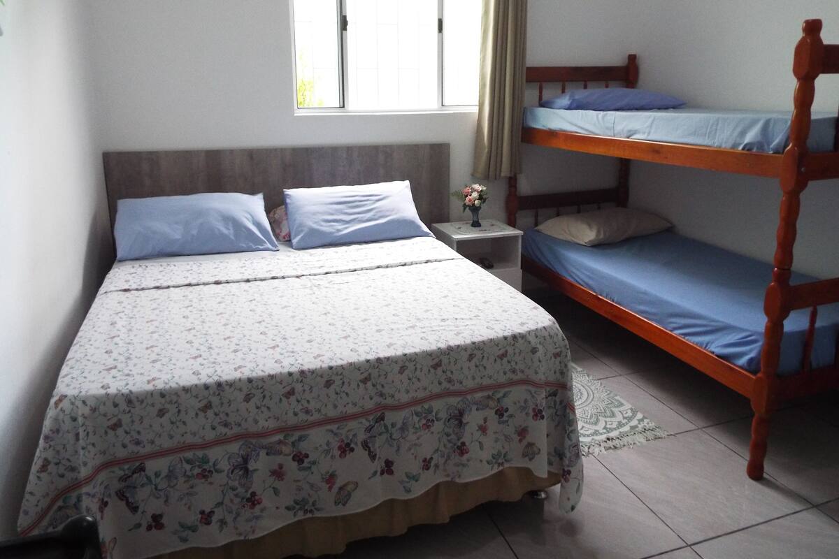 Une chambre avec un lit simple, un lit superposé, une table de chevet ornée d’un vase de fleurs et une fenêtre avec des rideaux.